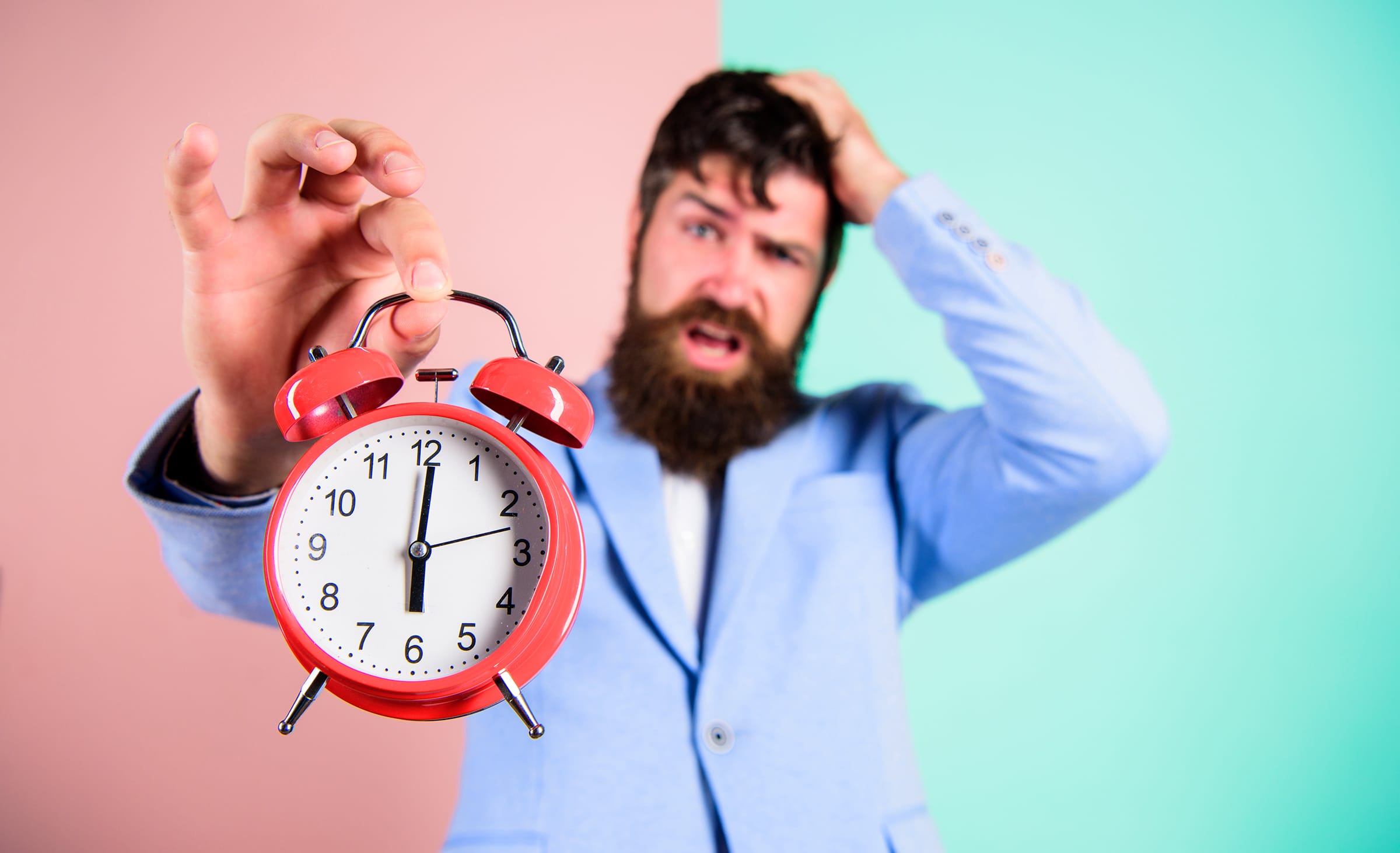 Bearded man in a blue shirt reaches toward the camera, holding a red alarm clock against a pink and mint background.