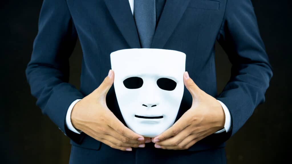 BusinessmanHideTheWhiteMaskInTheHandBehindHis | GroupOne Services Person in a navy suit holding a white mask in front of their chest, concealing their face to symbolize anonymity or deception.