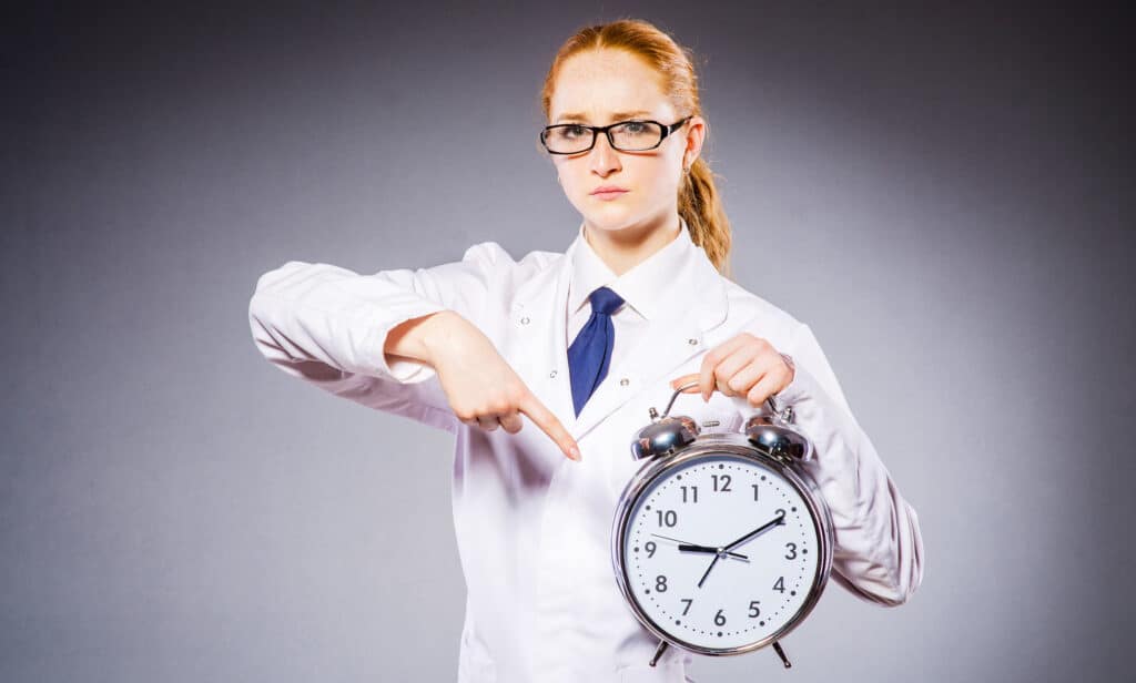 Woman in a lab coat holding a large alarm clock and pointing at it, suggesting a time reminder or deadline.
