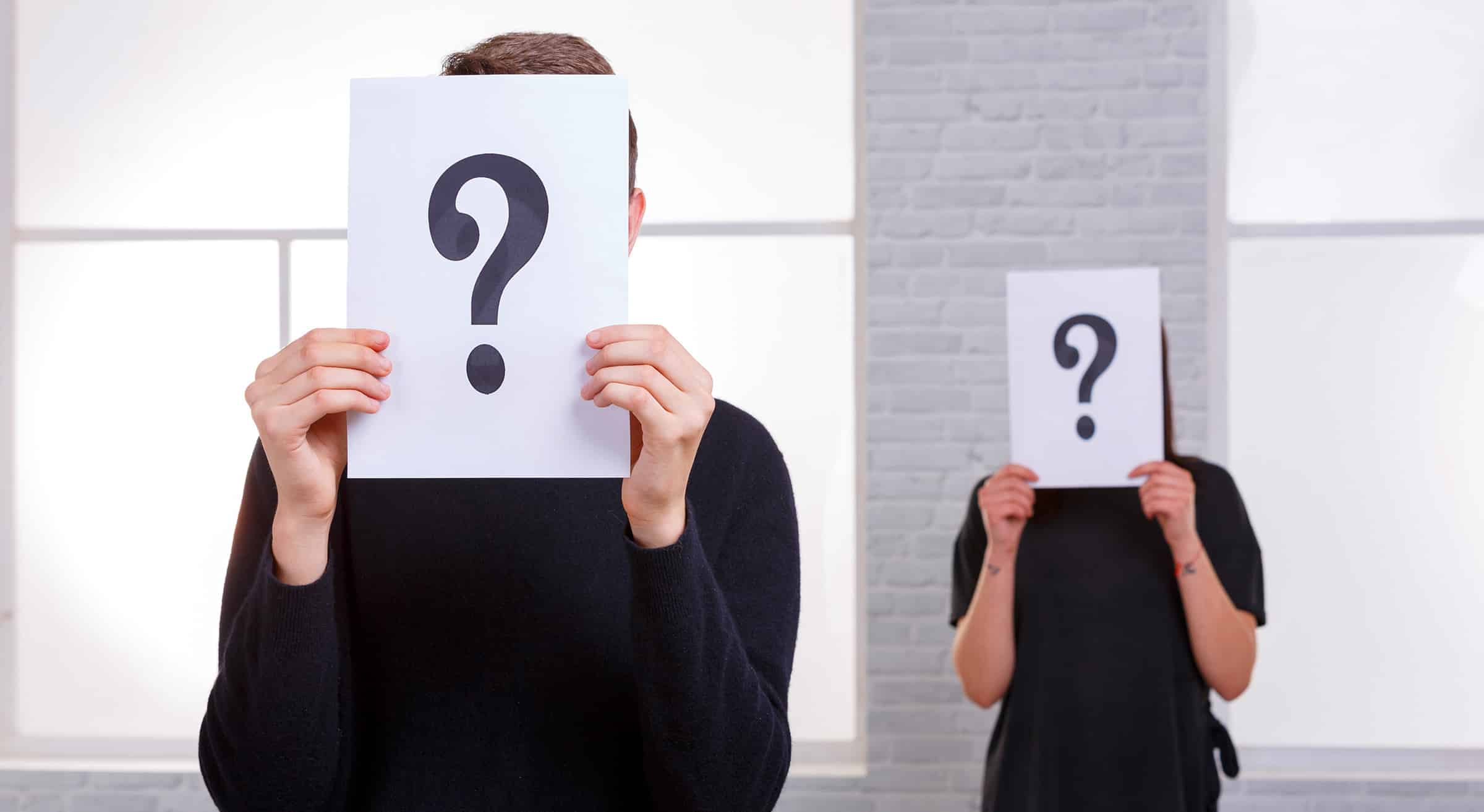 TwoPeopleCoveredTheirFacesWithAQuestionMark | GroupOne Services Two people in a bright room hold up white sheets with large question marks, obscuring their faces.