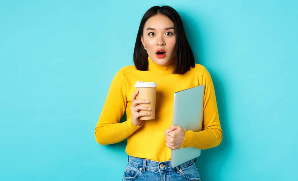 Young woman in a yellow sweater holds a coffee cup and a laptop, with a surprised expression against a turquoise background.