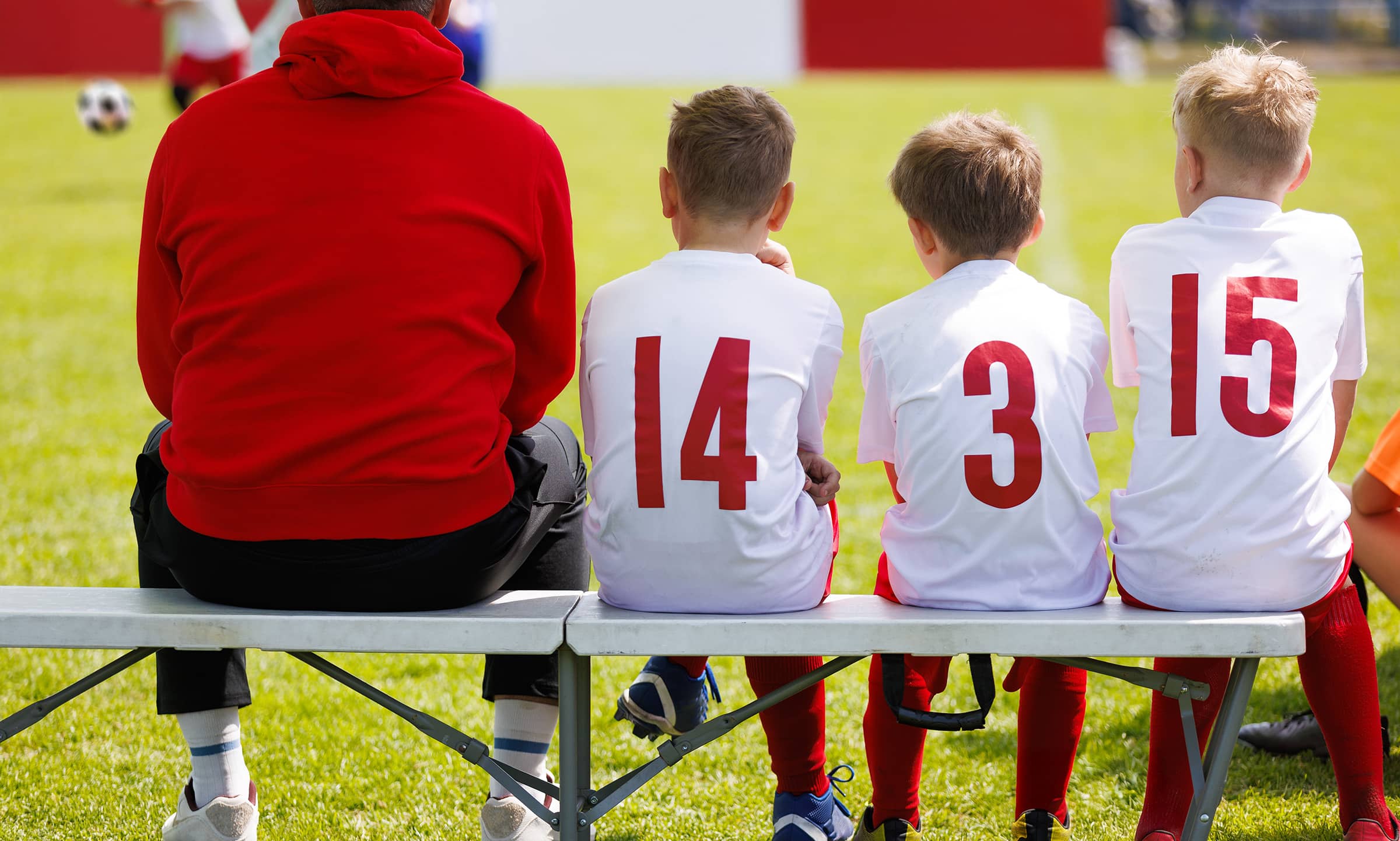 Kids in white soccer jerseys with red numbers sitting on a bench, facing a field while a coach in a red hoodie sits beside them on a sunny day.